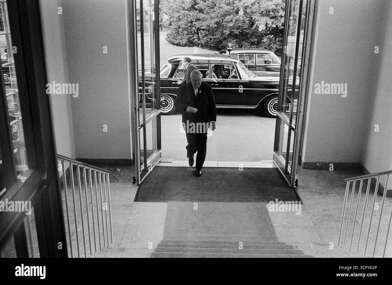 Ludwig Erhard betritt die offene Eingangstür eines Gebäude a Bonn. Draußen steht eine schwarze Staatslimousine des TYPS Mercedes-Benz 300 se (W112). Foto Stock