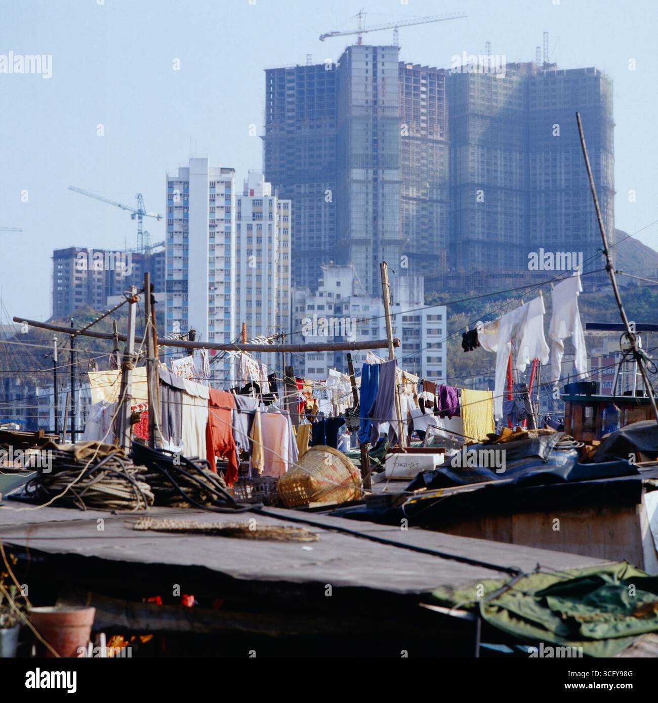 Auf einem schwimden Haus oder Boot hängt Wäsche zum Trocknen, im Hintergrund ragen Hochhäuser und Baukräne einer schnell wachsenden Stadt empor. Hong Kong, um 1980. Foto Stock