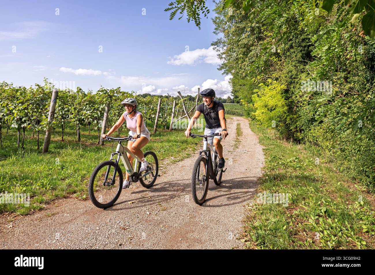 Coppia attiva che si gode un giro estivo in e-bike su un sentiero di ghiaia attraverso i vigneti della campagna slovena, uno stile di vita sano e il turismo all'aperto co Foto Stock