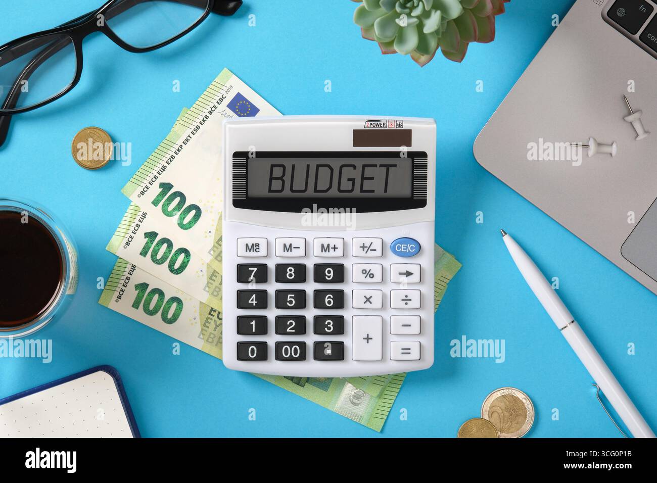 Calcolatore con testo di bilancio, banconote in euro, monete e forniture per ufficio su tavola blu. Pianificazione finanziaria e gestione del budget personale. Foto Stock