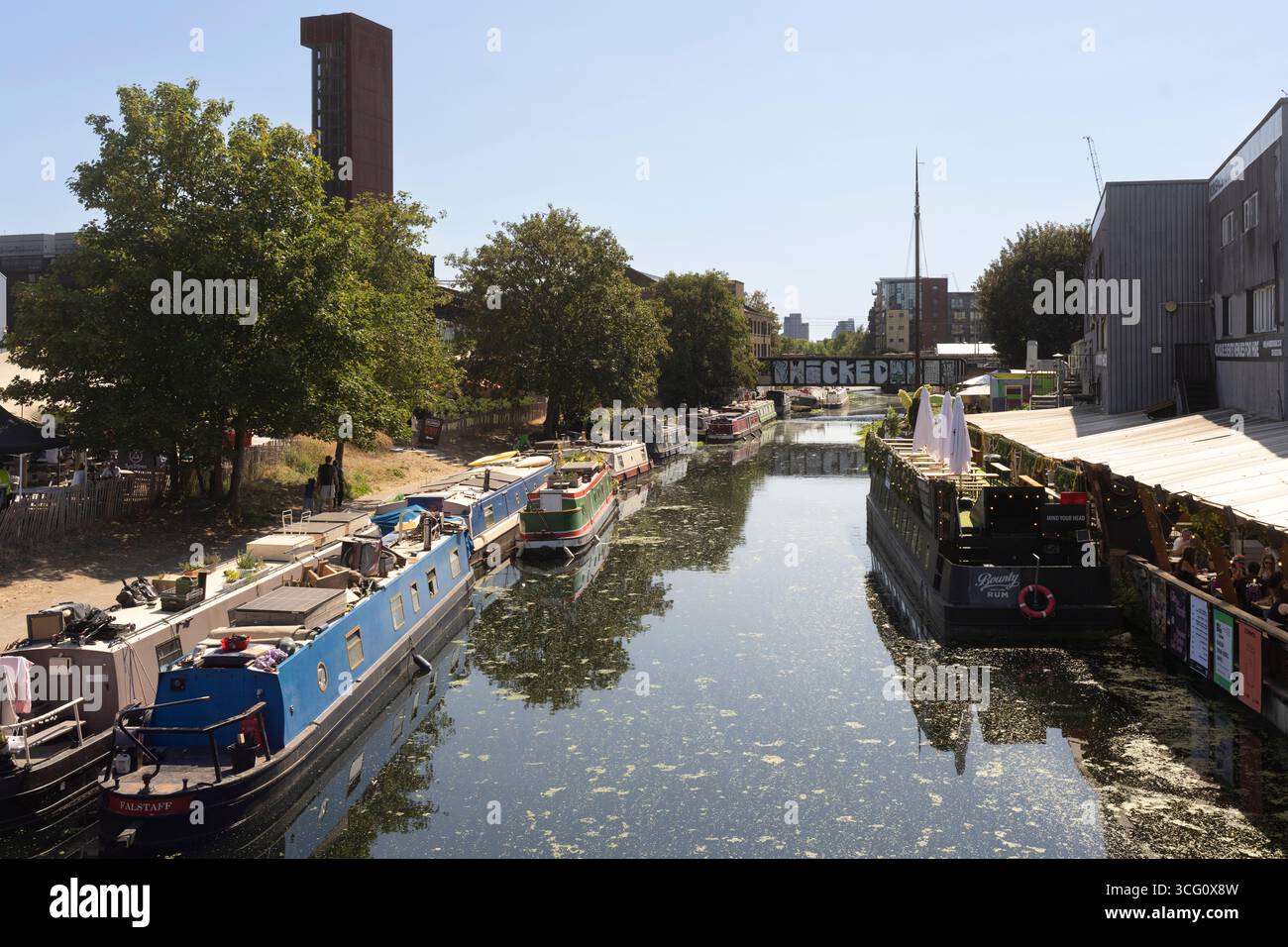 Le barche dei canali fiancheggiano il Regent's Canal a Hackney Wick, Londra. La gente ama mangiare e bere in una giornata di sole. Foto Stock