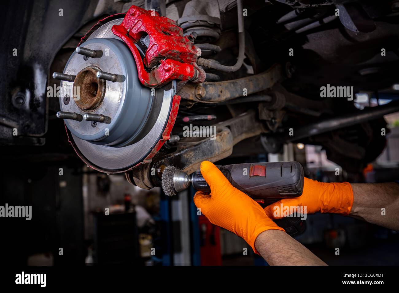 Un primo piano di un’auto sollevata in un garage, che mostra le mani di un lavoratore usando una macchina perforatrice per rimuovere la ruggine dai componenti arrugginiti di un supporto ruota, Highlig Foto Stock