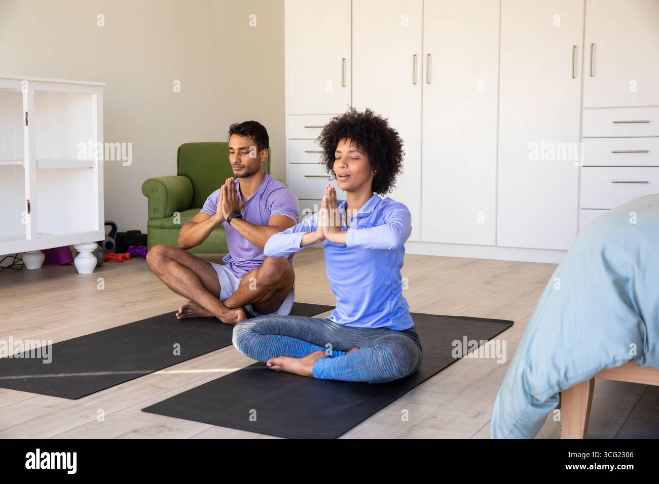 Coppie diverse che meditano su stuoie yoga in abbigliamento sportivo in camera da letto con armadietti e poltrona Foto Stock