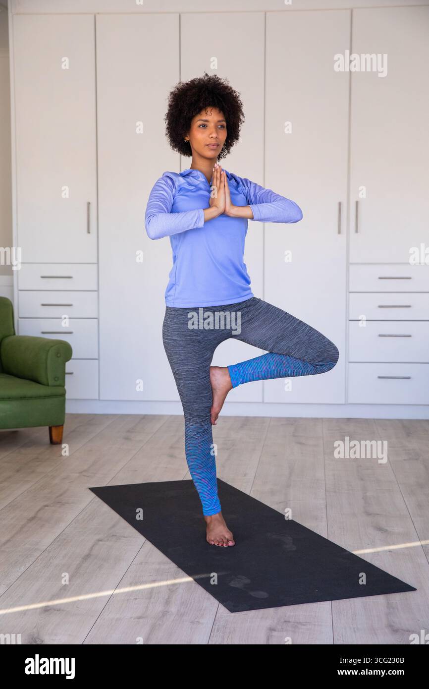 Donna afroamericana che tiene in posa l'albero su un tappetino da yoga nero nel soggiorno vicino alla poltrona Foto Stock