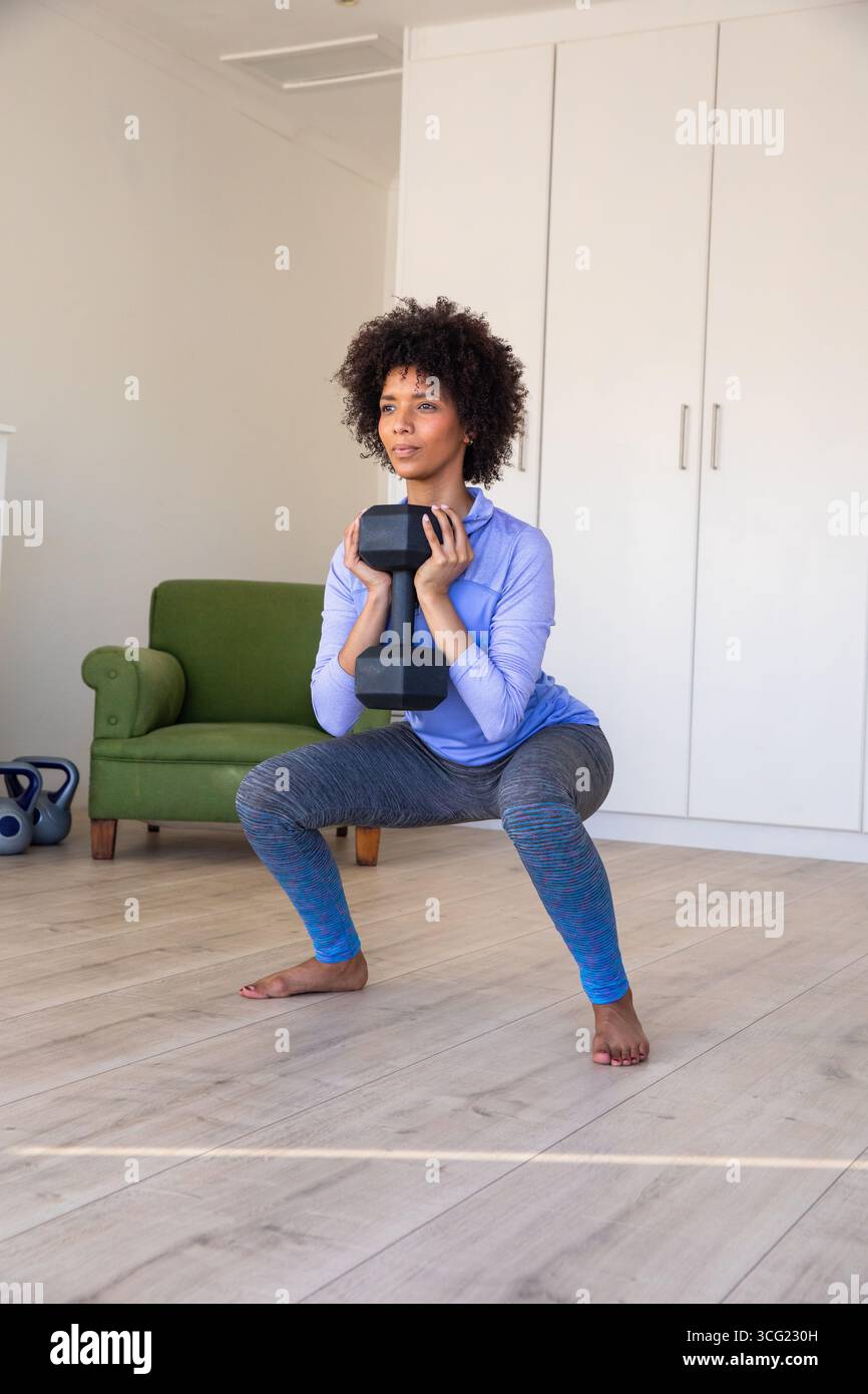 Donna afroamericana che esegue squat, stupida al petto in salotto con i kettlebell Foto Stock
