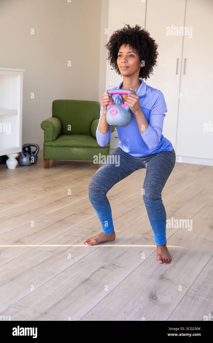 Donna afroamericana che accovacciata a casa indossa leggings con kettlebell grigio con manico rosa Foto Stock