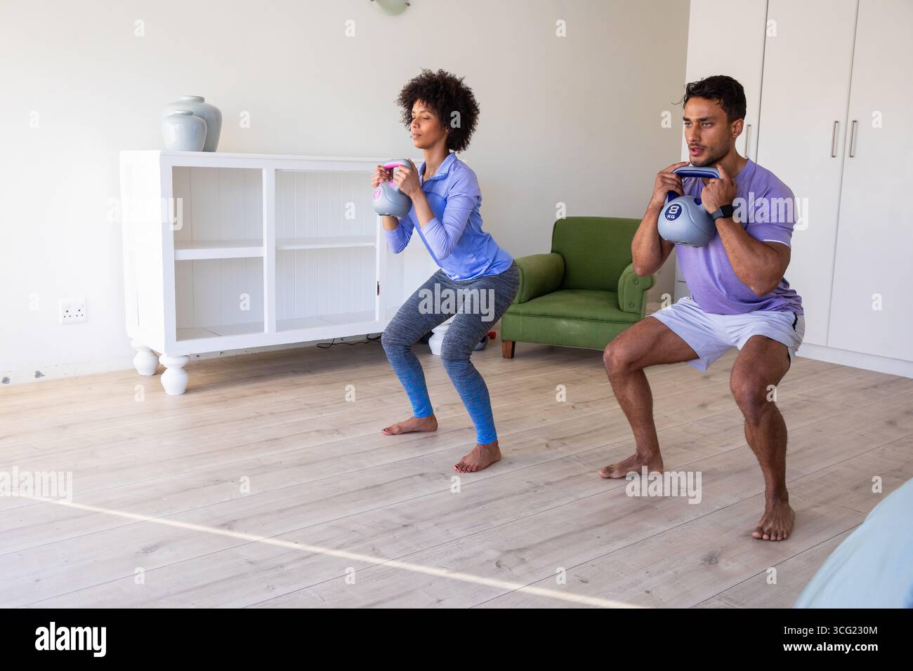 Coppia variegata con campanelli che eseguono squat nel soggiorno di casa vicino all'armadietto Foto Stock