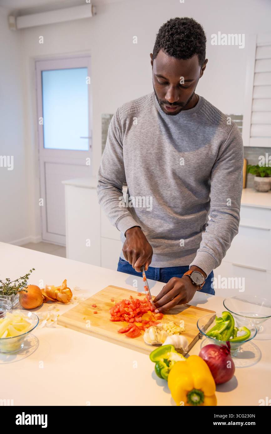 Uomo afroamericano di mezza età che taglia pomodori rossi su Kitchen Island con tagliere Foto Stock