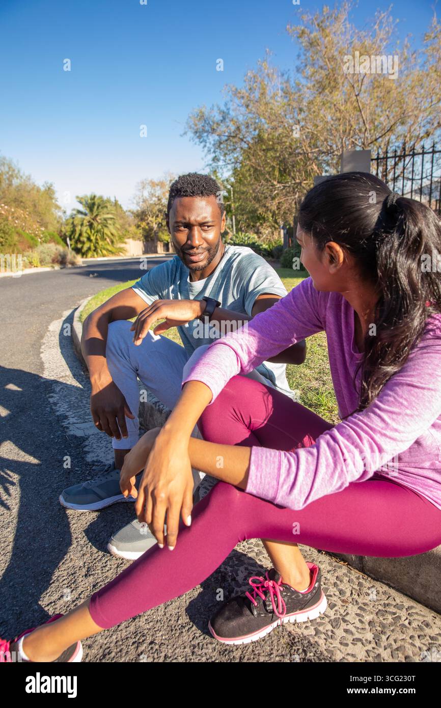 Diversi amici in abbigliamento sportivo seduti sul marciapiede suburbano per una pausa dopo una corsa Foto Stock