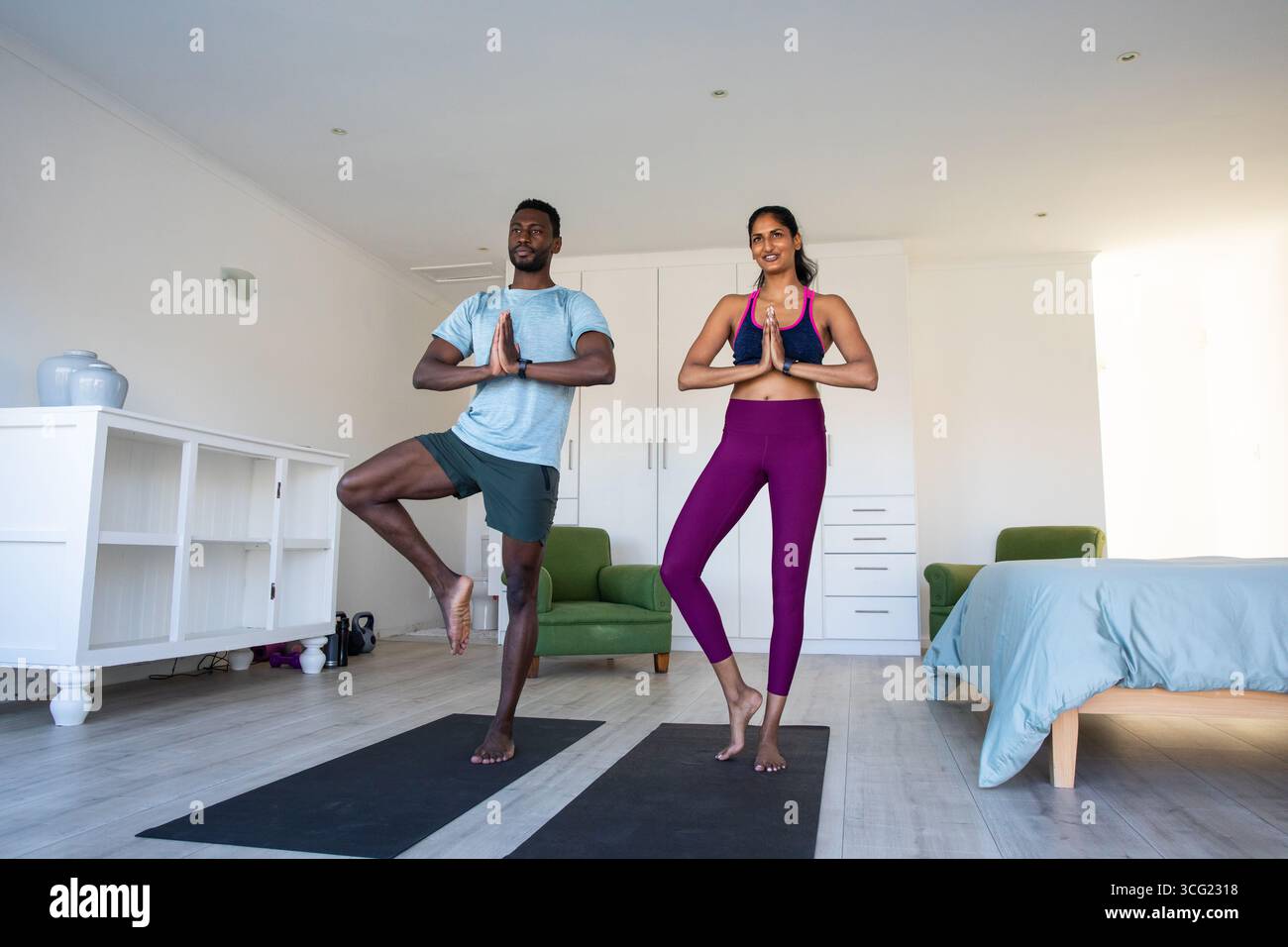 Coppie diverse che praticano lo yoga posano in camera da letto su tappetini da yoga con armadietti bianchi Foto Stock