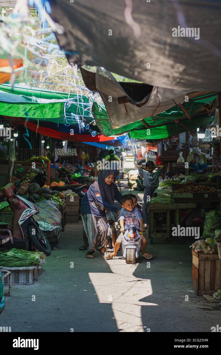 Balikpapan, Indonesia - 22 luglio 2025. Un momento di gioia mentre i bambini viaggiano in bici giocattolo attraverso un vivace mercato tradizionale, circondato da prodotti freschi e. Foto Stock
