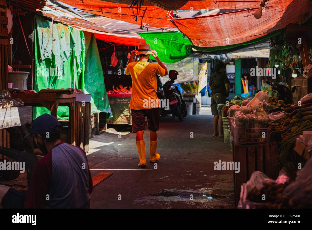 Balikpapan, Indonesia – 22 luglio 2025: Un uomo con una camicia e stivali giallo brillante si adatta al cappello mentre cammina attraverso un tradizionale vicolo rivestito di mercato Foto Stock
