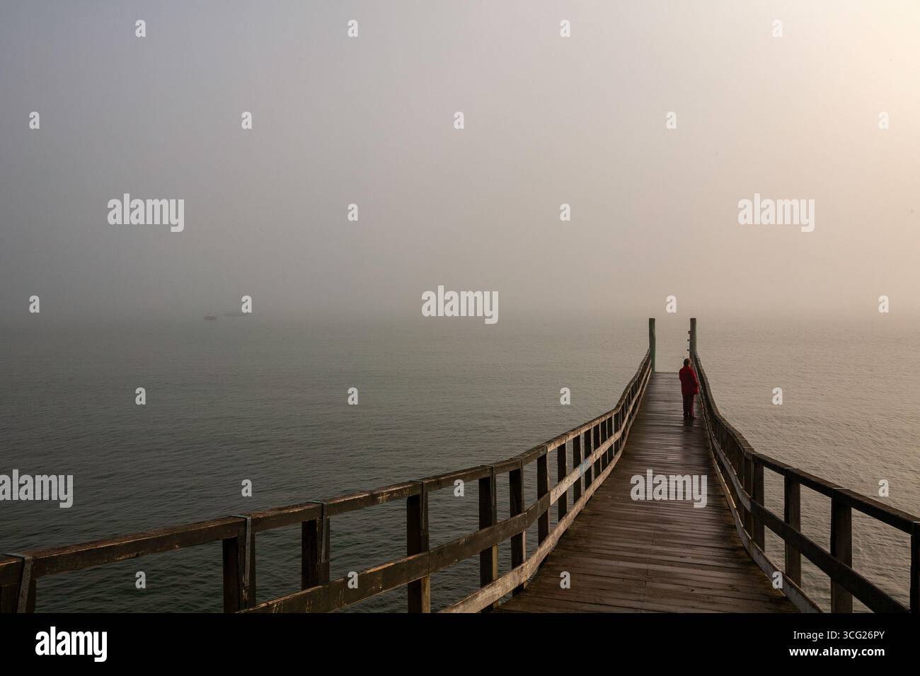 cappotto rosso donna in piedi sul lato destro del lungo molo molto pesante nebbia spessa nel porto sopra l'acqua luce solare luminosità da destra Foto Stock