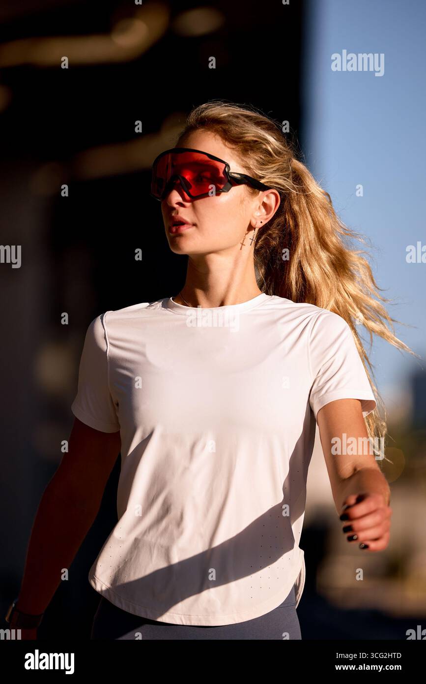 Una giovane donna fa jogging all'aperto, indossando occhiali da sole sportivi e abbigliamento sportivo. La luce del sole mette in risalto la sua espressione determinata mentre le piace Foto Stock