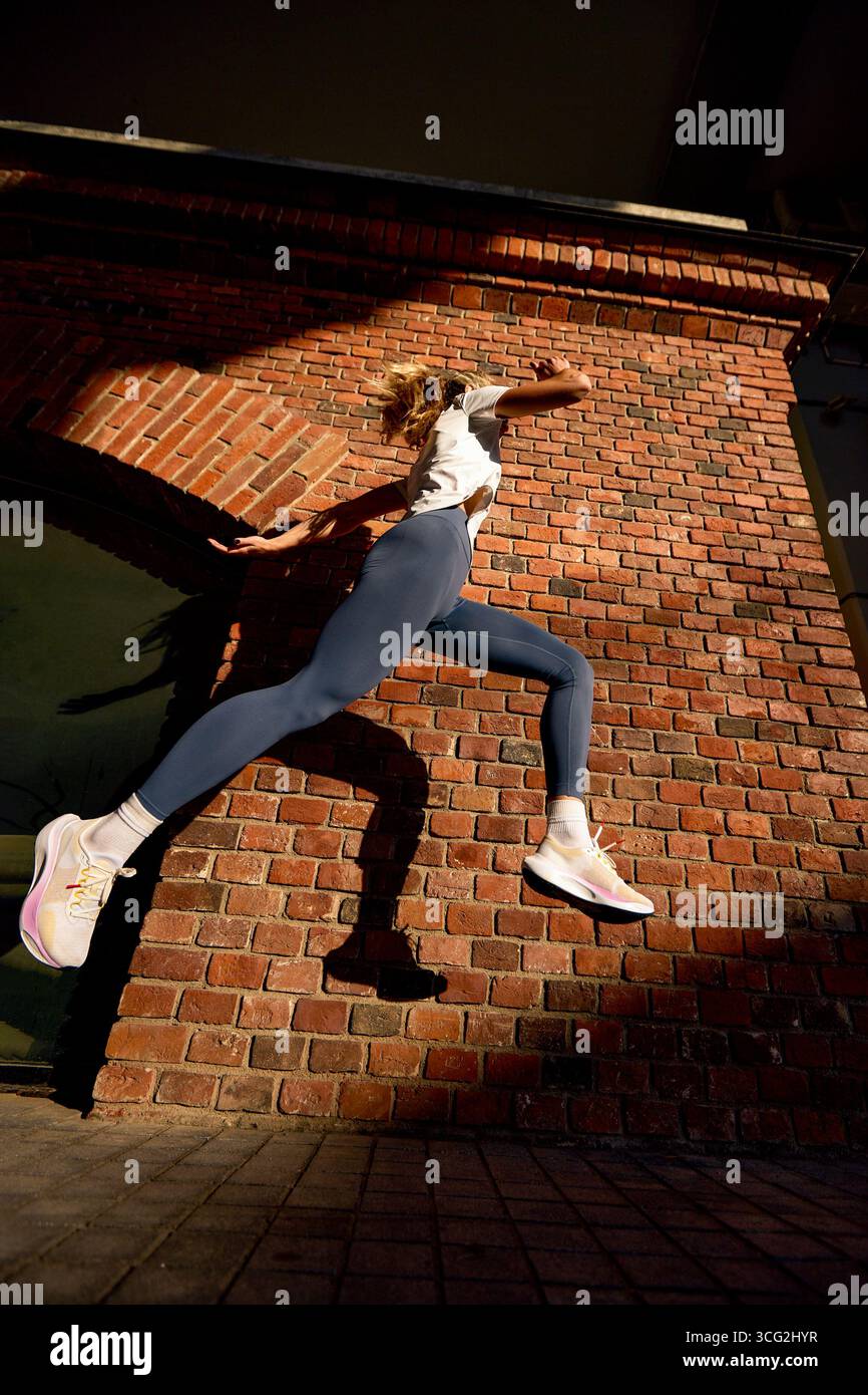 Una donna attiva viene catturata a metà salto su uno sfondo di mattoni testurizzati. La posa dinamica enfatizza il movimento e il fitness. La luce del sole proietta un look audace Foto Stock