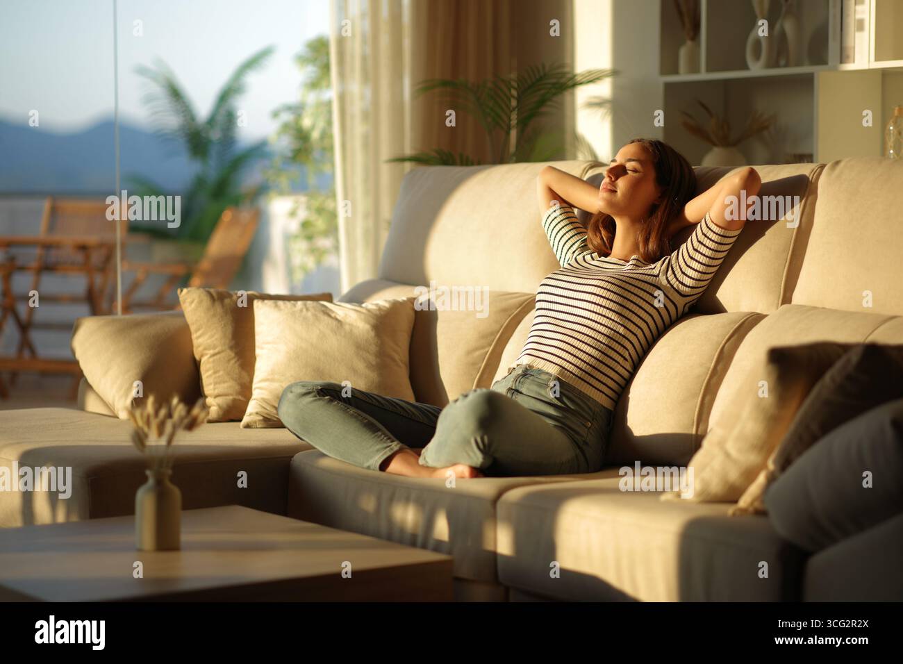 Donna rilassata seduta su un comodo divano nel soggiorno al tramonto a casa Foto Stock