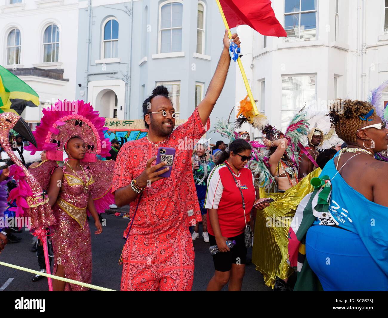 L'uomo in abito arancione con occhiali insoliti regge una bandiera durante il Carnevale di Notting Hill, Londra. Inghilterra 25 agosto 2025 Foto Stock