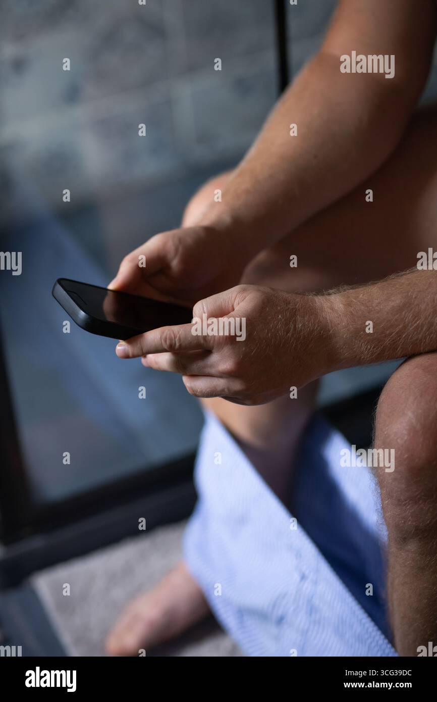 Uomo caucasico seduto sulla ciotola del WC in bagno usando il suo smartphone. Dipendenza, dipendenza dai social network. Foto Stock