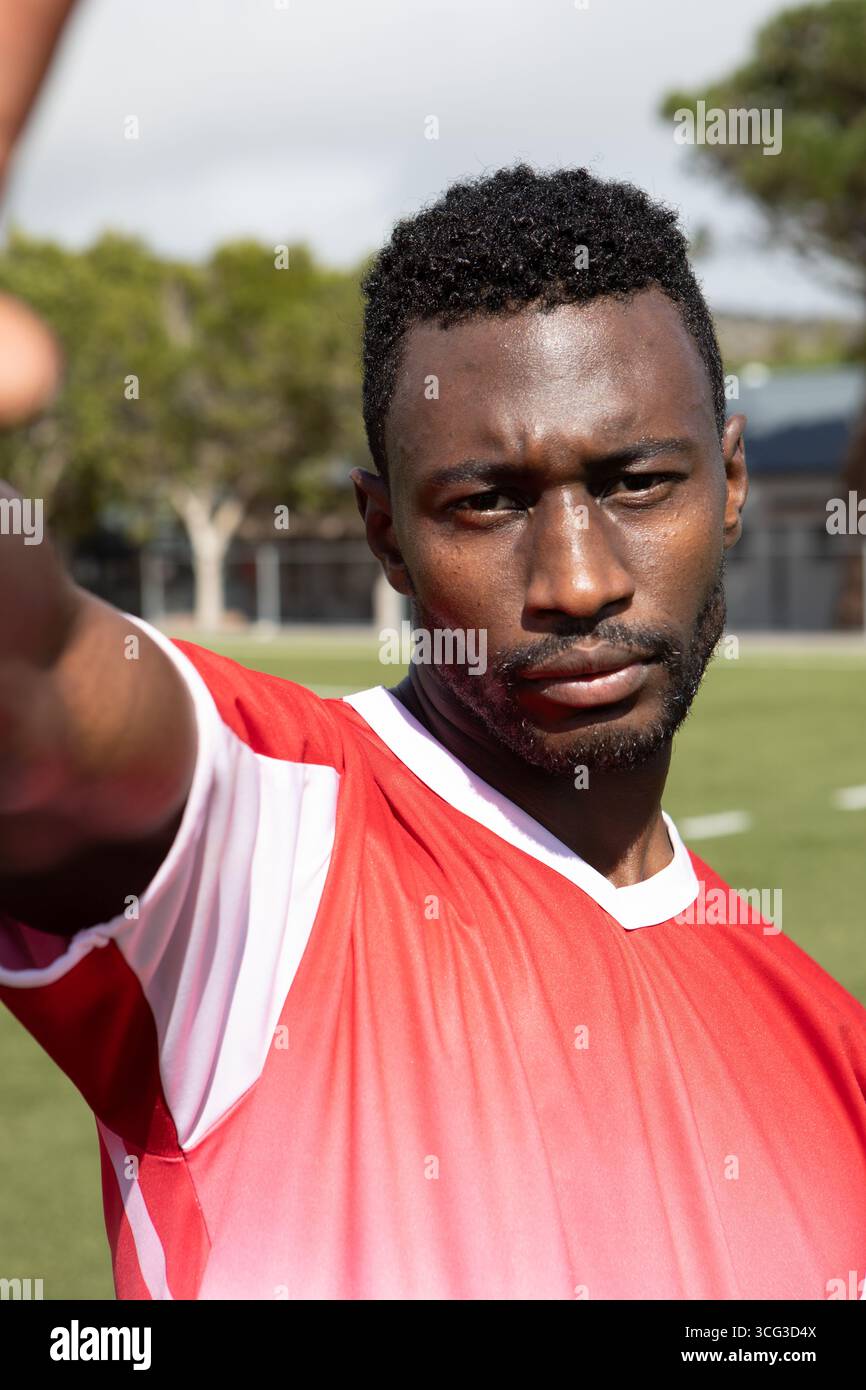 Uomo afroamericano in piedi sul campo sportivo allungando il braccio alla telecamera indossando una maglia rossa e bianca Foto Stock