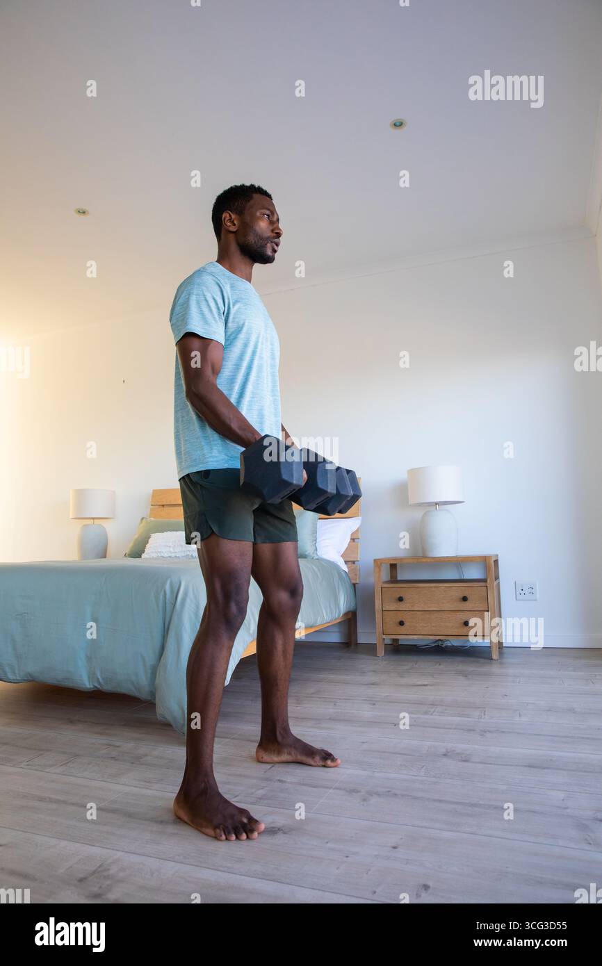 Uomo afroamericano che solleva manubri esagonali in una camera da letto ordinata accanto al letto di legno, lampade Foto Stock
