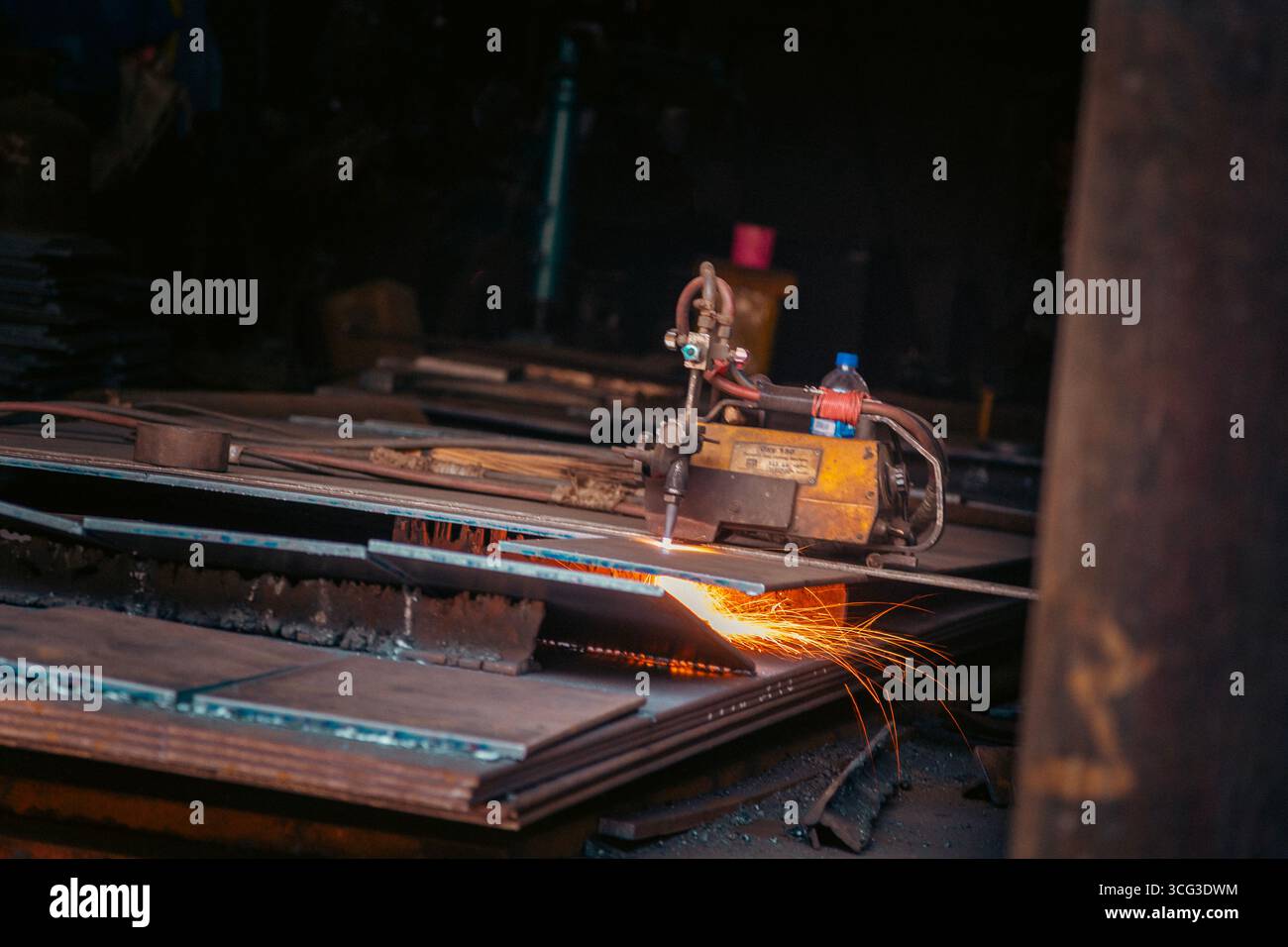 Questa immagine cattura una torcia da taglio industriale che affetta lamiere, con scintille luminose e macchinari in un ambiente di officina Foto Stock