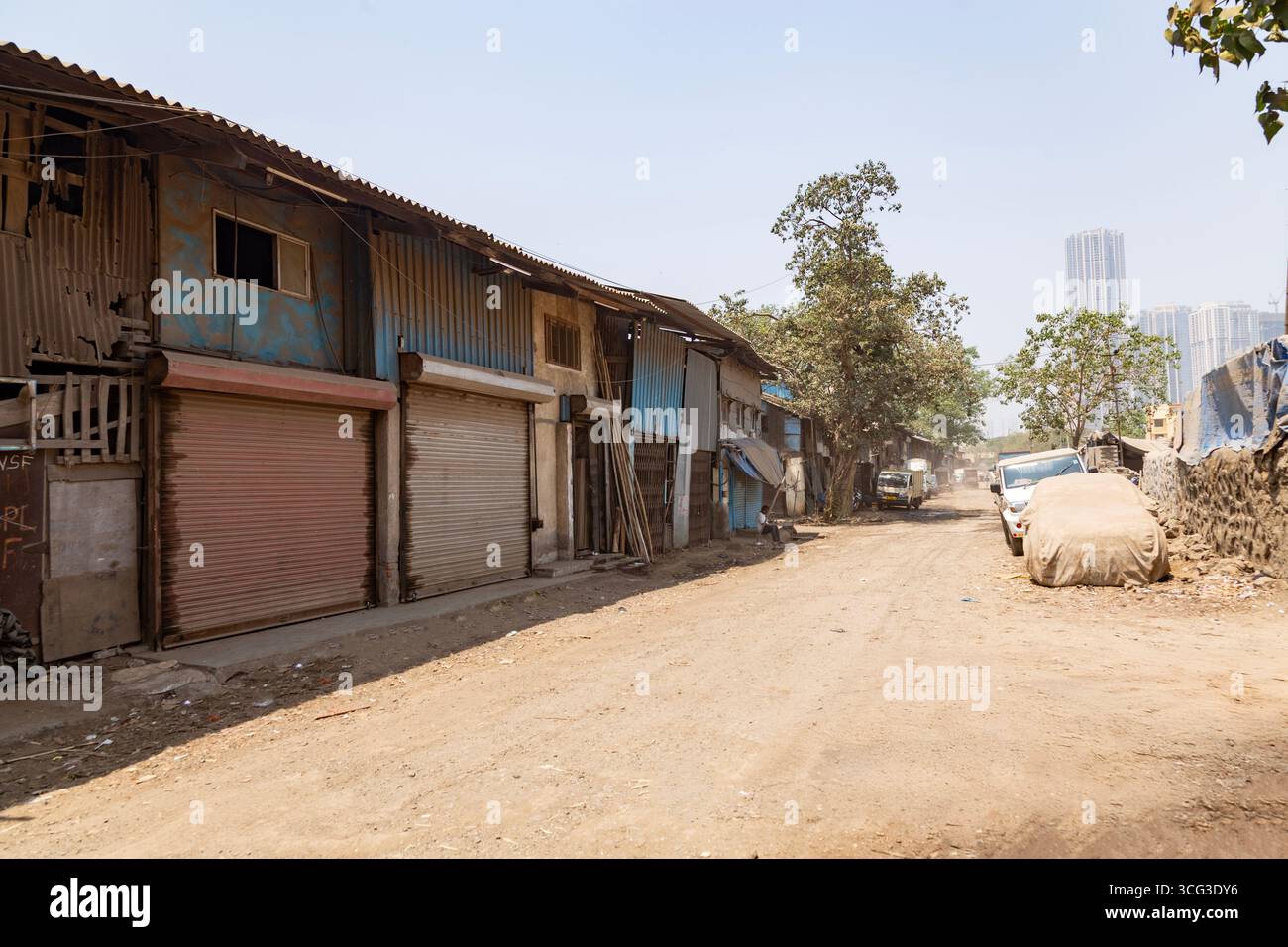 Questa immagine mostra una strada polverosa fiancheggiata da baracche in metallo e legno, persiane chiuse, un albero e veicoli parcheggiati, catturando la povertà urbana Foto Stock
