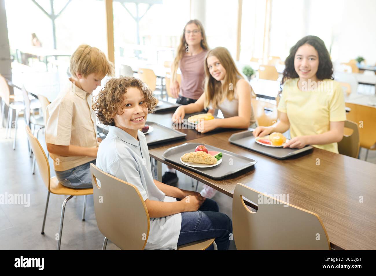 Un gruppo di bambini allegri seduti in una luminosa caffetteria scolastica, che si gusta i pasti insieme. Cibo fresco e sano su vassoi crea un'atmosfera vivace Foto Stock