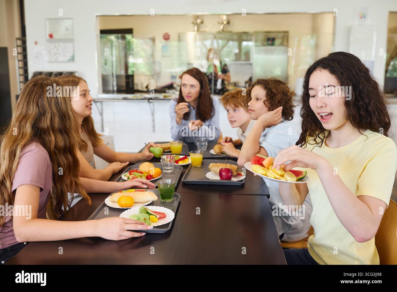 Un gruppo di studenti sta mangiando vari pasti freschi e salutari e si sta godendo la conversazione in una caffetteria scolastica ben illuminata e pulita. L'atmosfera e' vivace Foto Stock