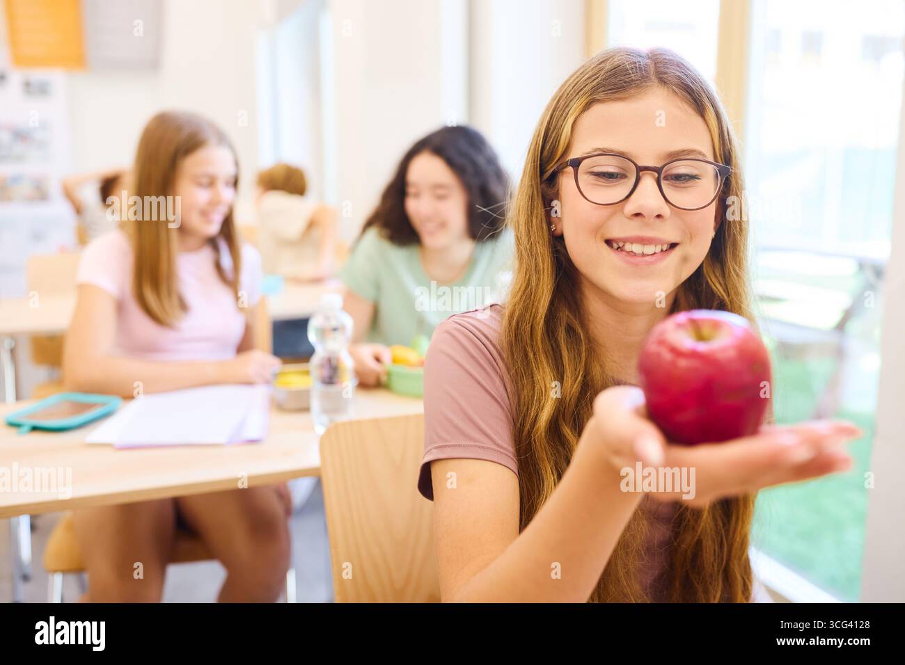 Un ambiente scolastico allegro con gli studenti che condividono un momento durante lo spuntino, concentrandosi su uno studente che ammira una mela rossa fresca in una classe ben illuminata Foto Stock