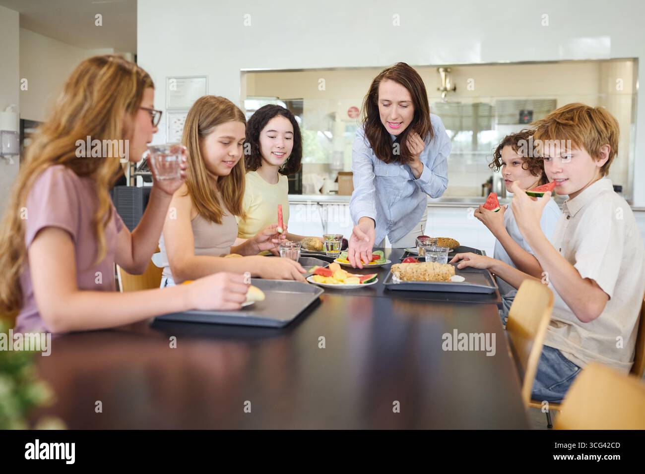 Un gruppo di studenti e un adulto si riuniscono in una caffetteria ben illuminata, condividendo il pranzo. La scena cattura un momento di comunità, alimentazione sana e studente Foto Stock