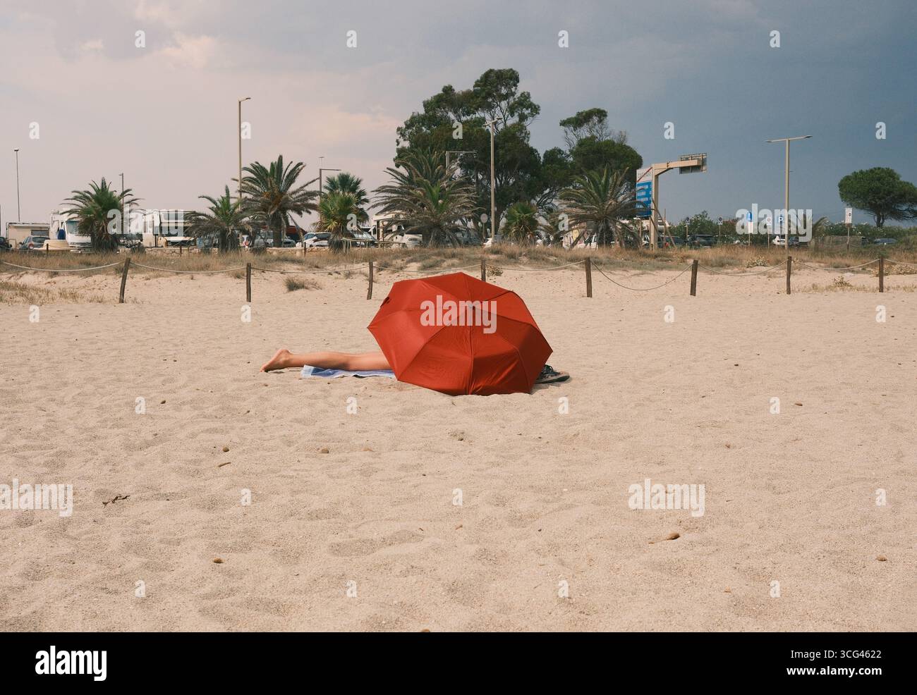 Una tranquilla spiaggia caratterizzata da un vibrante ombrello rosso che ombrega un sole su soffice sabbia dorata, circondato da lussureggianti palme e da un cielo nuvoloso. Foto Stock