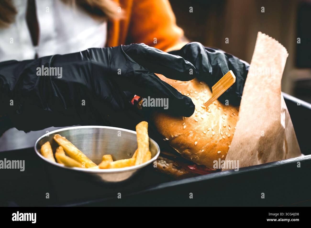 Mani femminili in guanti neri che reggono deliziosi hamburger freschi con patatine fritte sul tavolo di legno nero Foto Stock