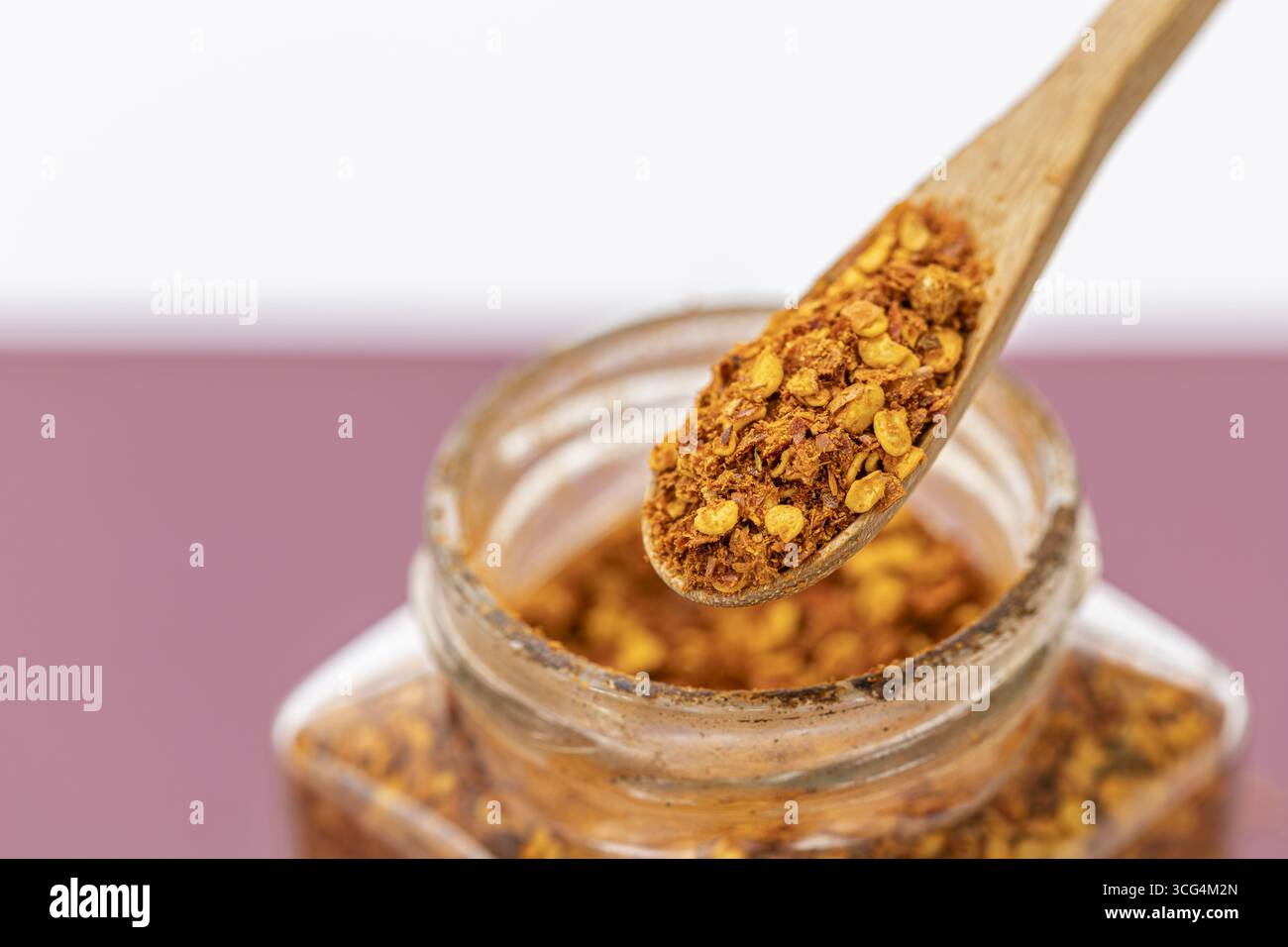 Un cucchiaio di legno con fiocchi di peperoncino vaso di vetro per condimenti piccanti primo piano ingrediente da cucina di colore rosso aromatico che esalta lo stile rustico Foto Stock