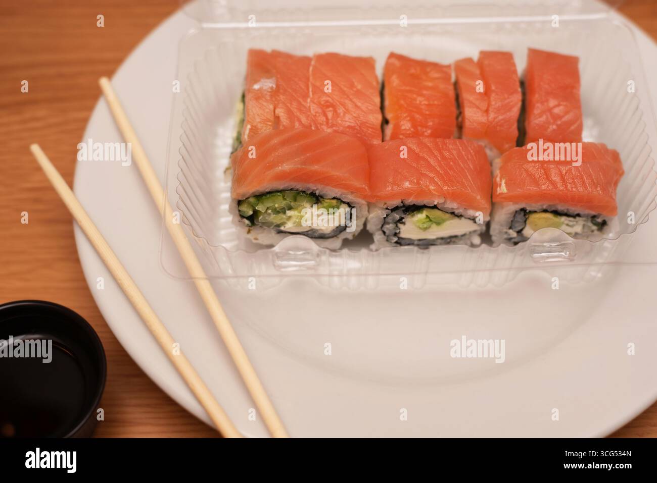 Panini di sushi con salmone fresco, avocado e formaggio spalmabile serviti in un contenitore di plastica trasparente sul piatto, accompagnati da bacchette e salsa di soia. Un'app Foto Stock