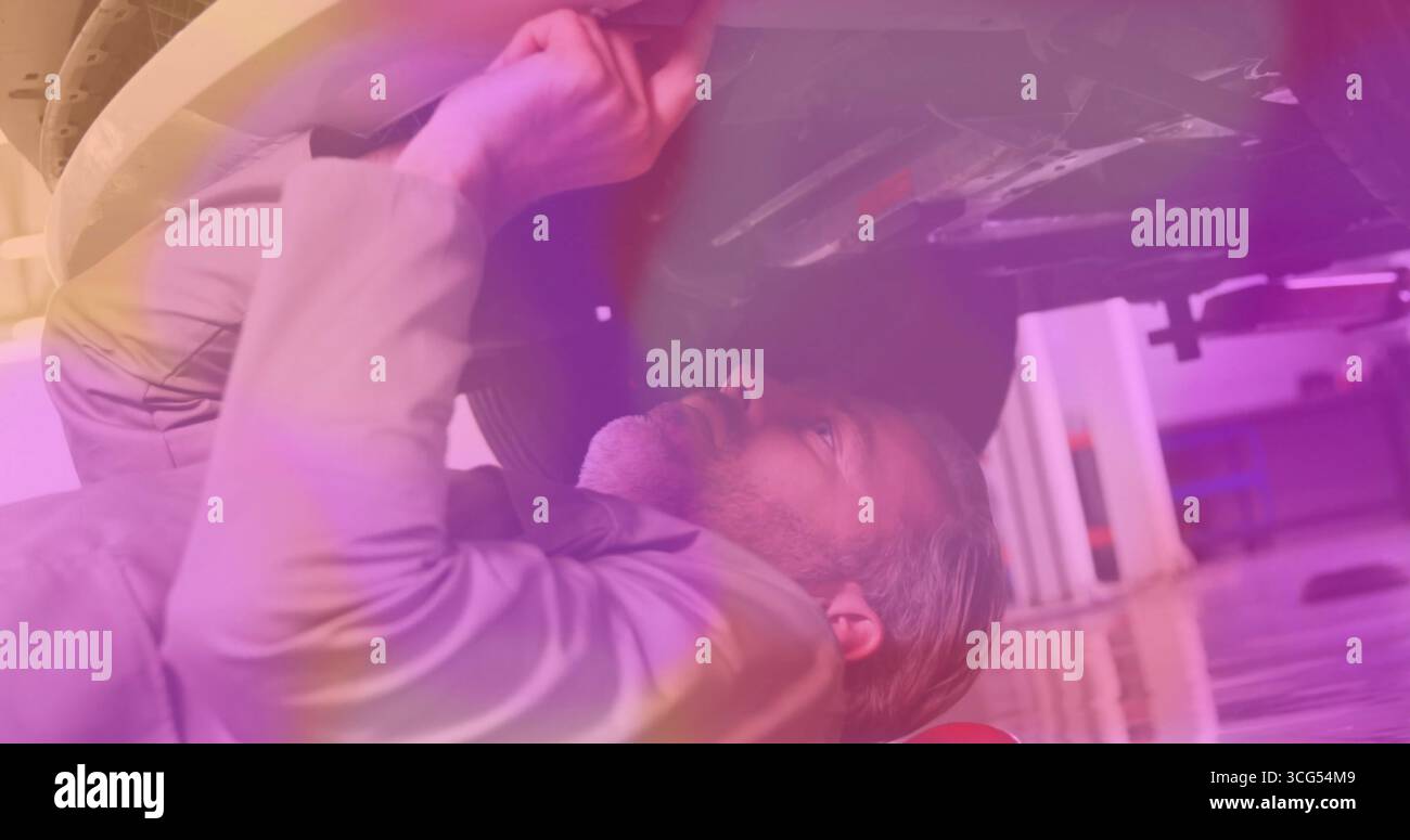 Meccanico senior che indossa una camicia da lavoro giacente sotto il veicolo in garage, con pneumatici e poggiatesta superriduttore Foto Stock