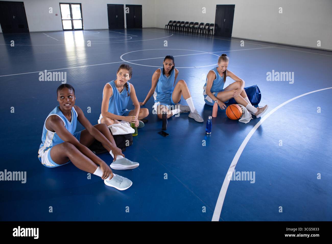 Diverse giocatrici di basket che riposano sul campo dal basket, adattano le sneakers e sorseggiano acqua Foto Stock