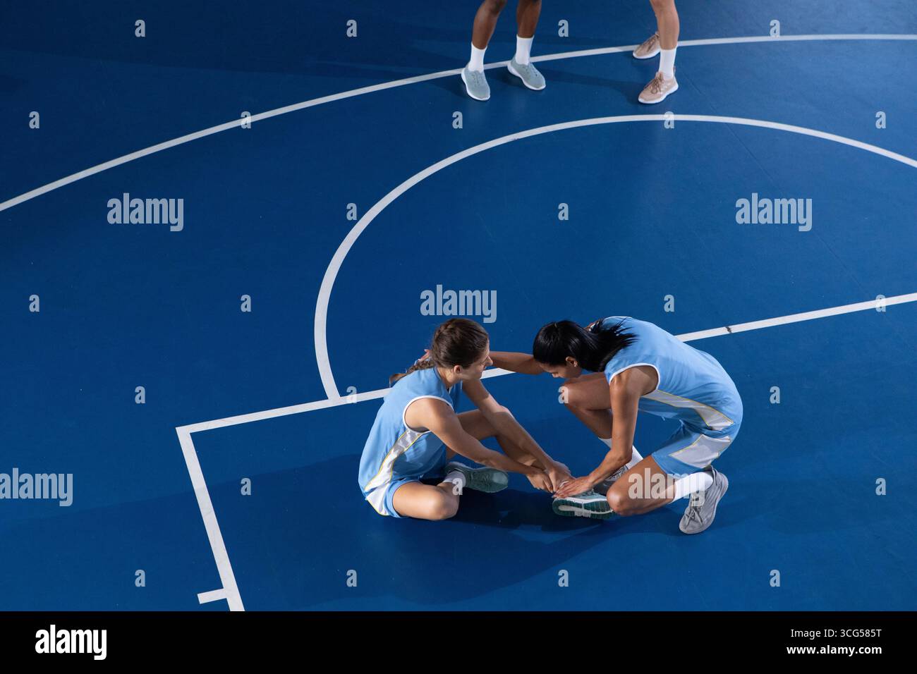 Diverse compagne di squadra di basket si allungano in avanti mentre si siedono sul campo blu indossando uniformi Foto Stock