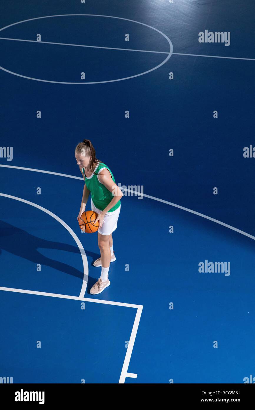 Giocatrice di basket in maglia verde che regge il basket e si trova sul campo blu sotto le luci Foto Stock