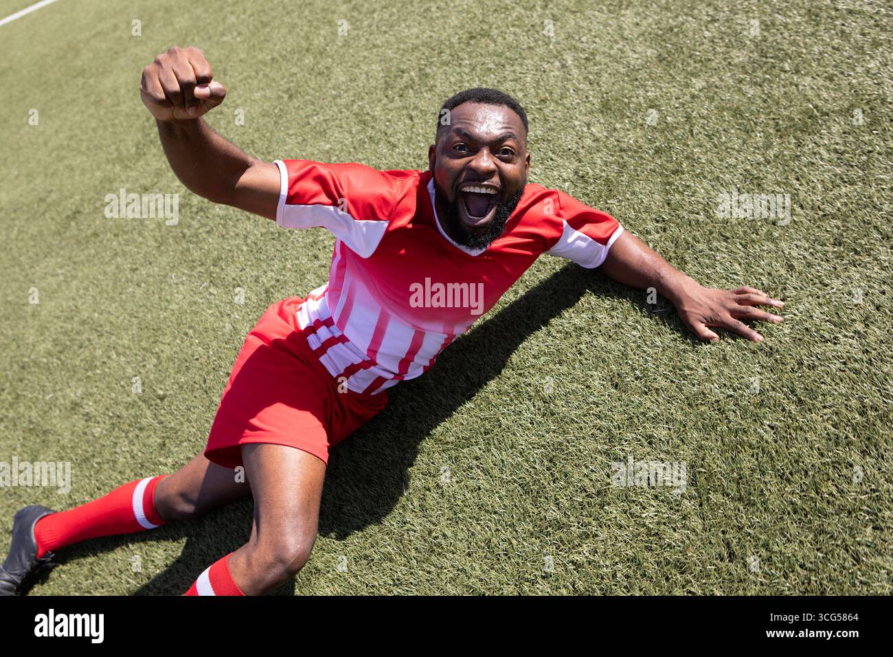 Giocatore di calcio afroamericano che scivola sul prato celebrando il gol indossando scarpe rosse Foto Stock