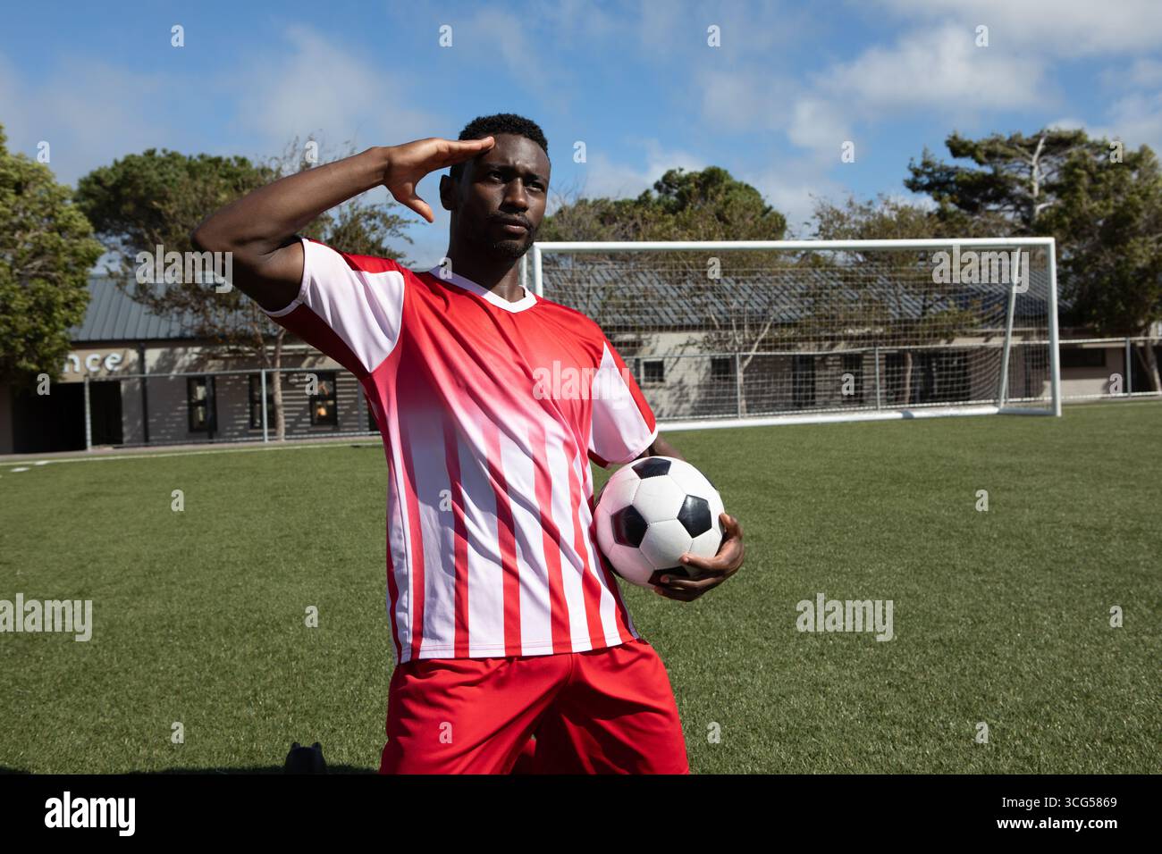 Giocatore di calcio afroamericano che saluta e tiene la palla in porta in maglia rossa e pantaloncini corti Foto Stock
