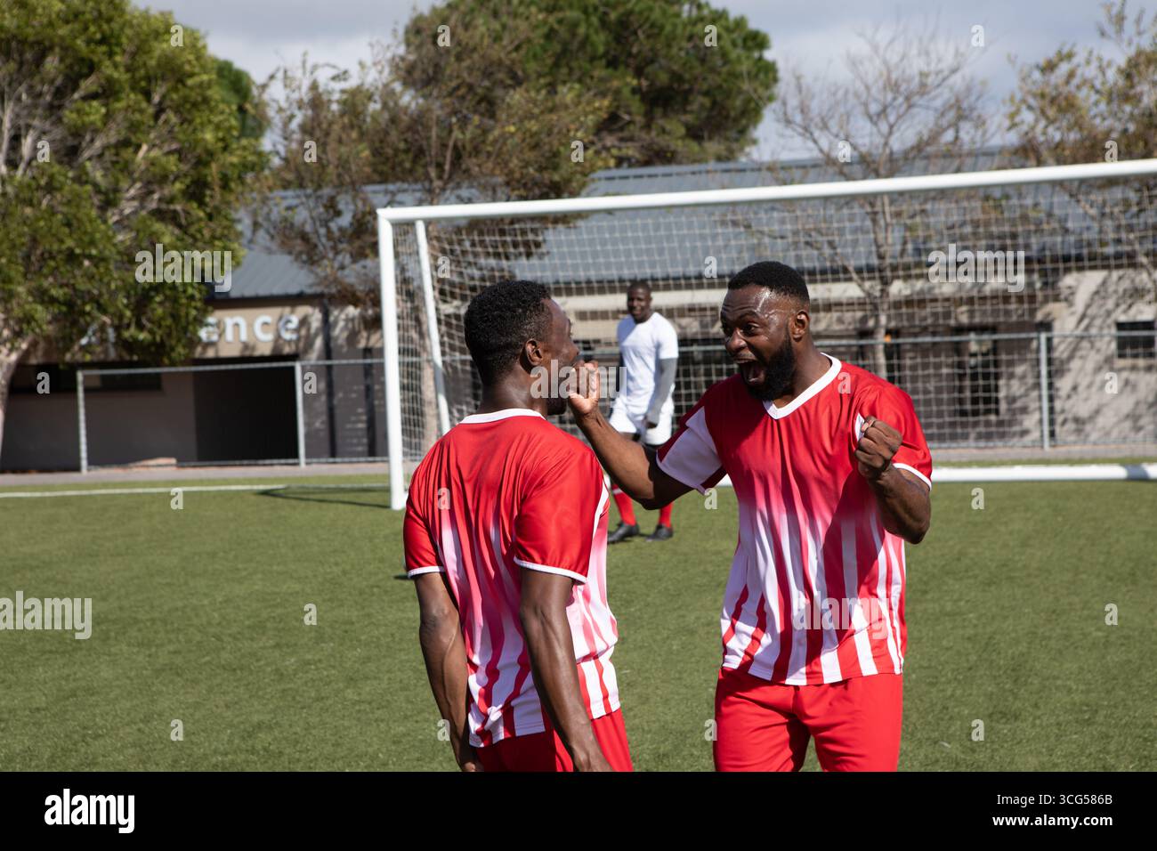 Compagni di squadra afroamericani che celebrano il gol in divise rosse e bianche sul campo da calcio vicino alla rete Foto Stock