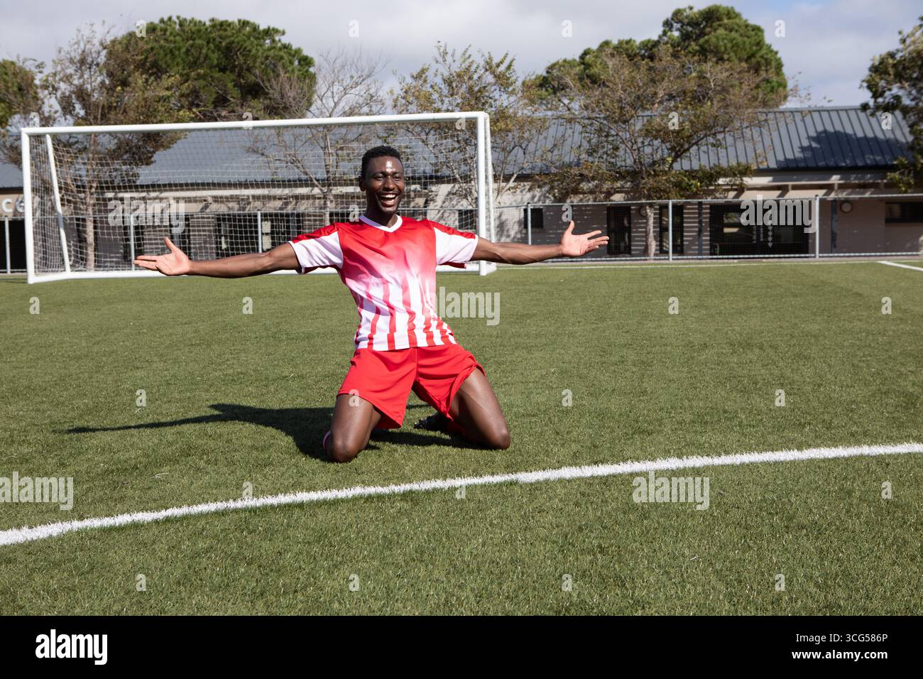 Uomo afro-americano che indossa una maglia inginocchiata sul campo di prato a porta di calcio tifa con le braccia alzate Foto Stock