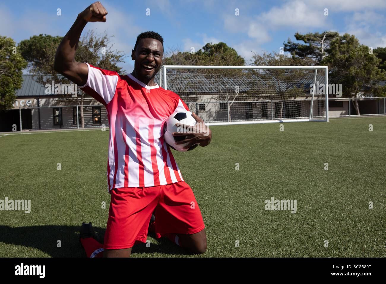 Uomo afroamericano in maglia rossa inginocchiato sul campo da calcio, con palla in porta, pugno di pompaggio Foto Stock