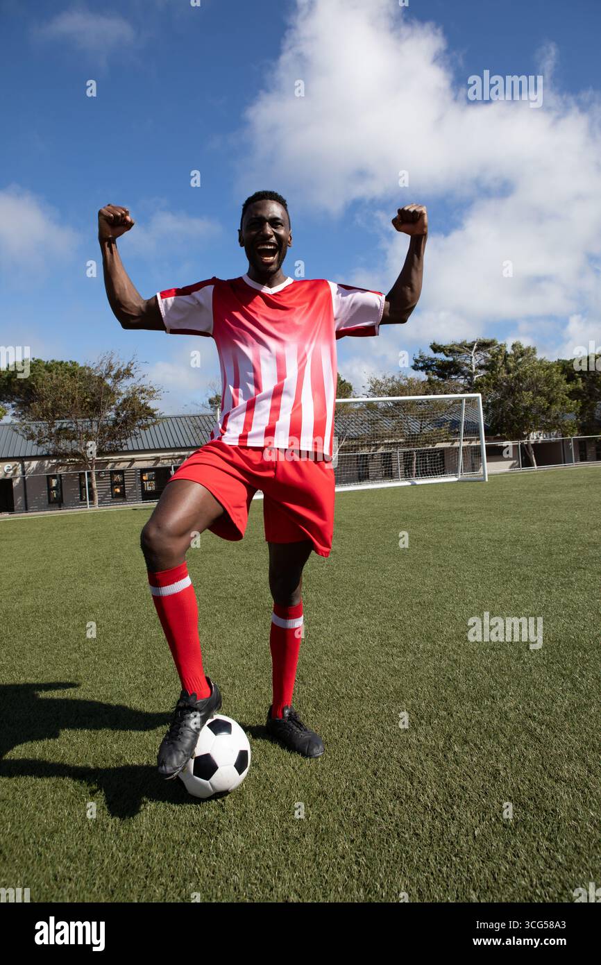 Uomo afroamericano che festeggia con un piede sul pallone da calcio sul campo artificiale in erba vicino alla rete porta Foto Stock