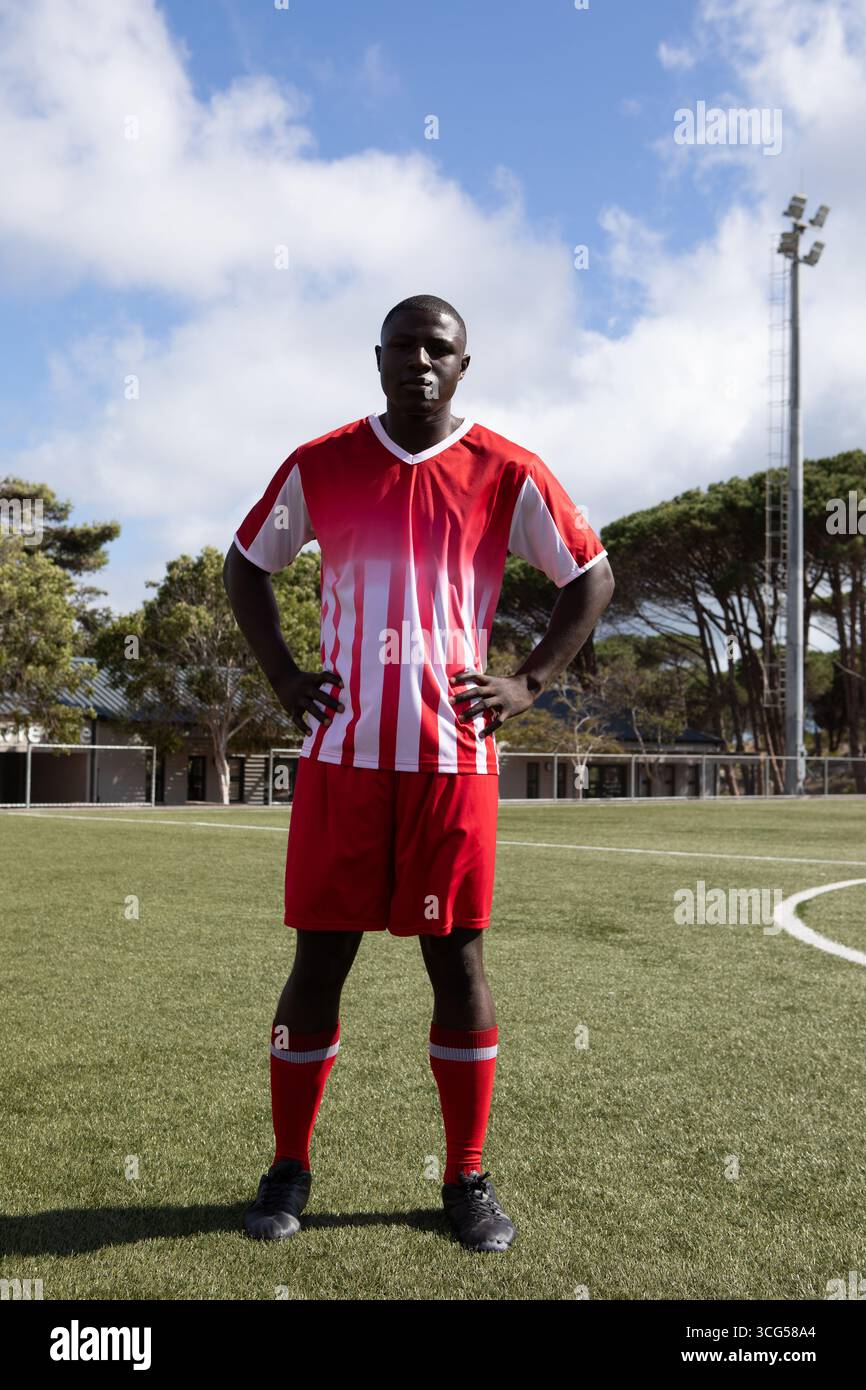 Giocatore di calcio afroamericano maschile in piedi sul campo da calcio in erba con torre a riflettore in divisa rossa Foto Stock