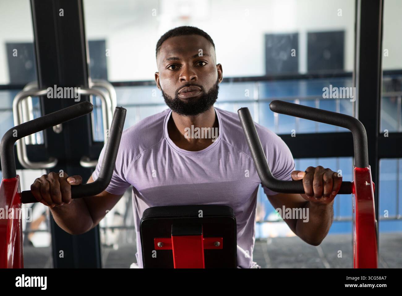 Uomo afro-americano che spinge i manici del petto nella zona pesi della palestra indossando una T-shirt viola leggera Foto Stock