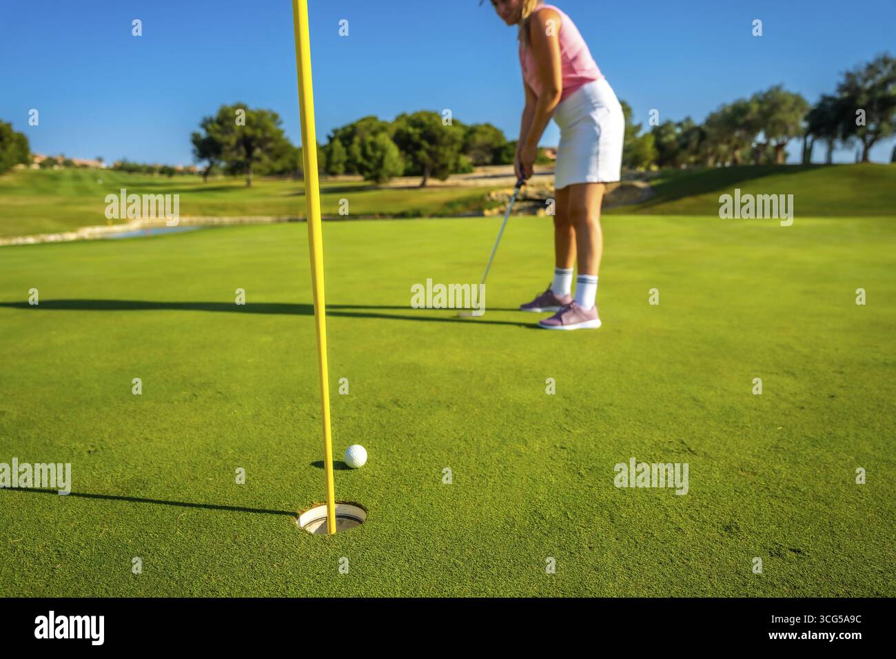 Donna che mette abilmente su un campo da golf, puntando verso la buca sotto un cielo azzurro. I lussureggianti dintorni verdi accentuano l'atmosfera serena Foto Stock