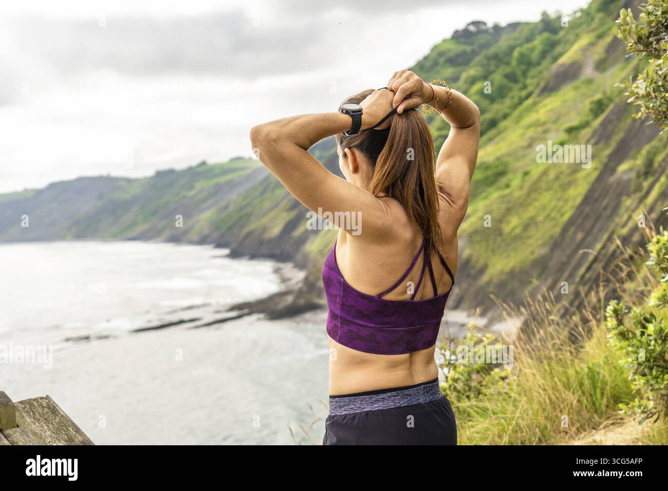 Vista posteriore di una donna sportiva che lega la sua coda di cavallo mentre si gode la vista del mare e delle scogliere flysch sulla spiaggia di sakoneta a zumaia, campagna basca, sp Foto Stock