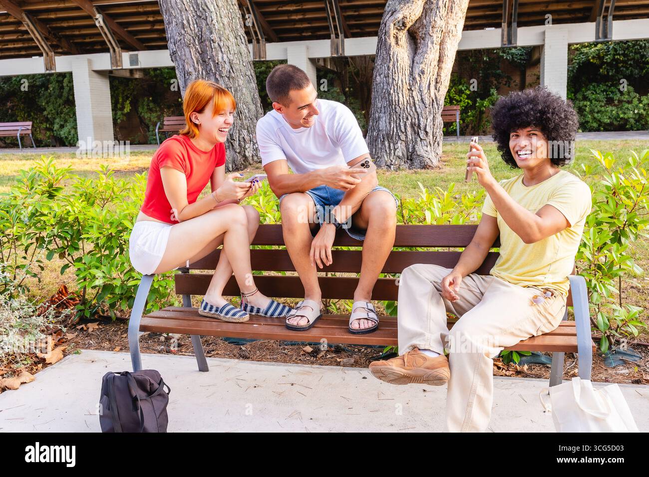 Giovani amici che usano smartphone e ridono insieme su una panchina in un parco Foto Stock