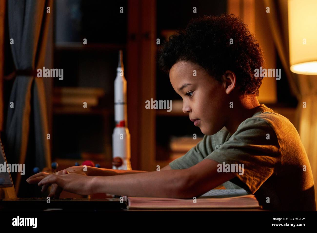 Vista laterale del ragazzo Alpha Black seduto alla scrivania di fronte al portatile nel soggiorno di casa in cerca di informazioni sui pianeti Foto Stock
